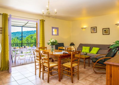 Salon d'un appartement avec espace repas, un canapé, et deux fauteuils noirs. En arrière-plan, terrasse avec table dressée pour le petit-déjeuner