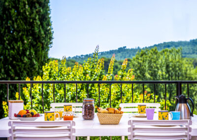 Sur la terrasse d'un appartement, table préparée pour le petit-déjeuner avec céréales, viennoiseries, café et fruits, offrant une vue panoramique sur la nature environnante