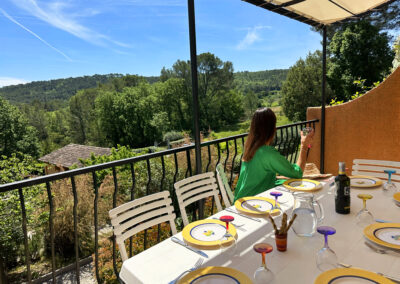 Table à manger dressée pour un repas sur la terrasse d'un appartement, où une femme tient un verre en appréciant la vue panoramique