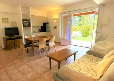 Salon avec canapé confortable, table basse, espace repas, télévision, et cuisine entièrement équipée. En arrière plan, jardin comme terrasse au rez-de-chaussée offrant une vue agréable sur l'extérieur